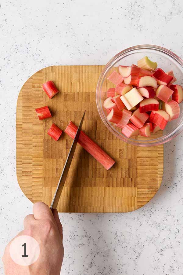 A hand slicing rhubarb stalks into ¾-inch pieces on a wooden board for vodka infusion.