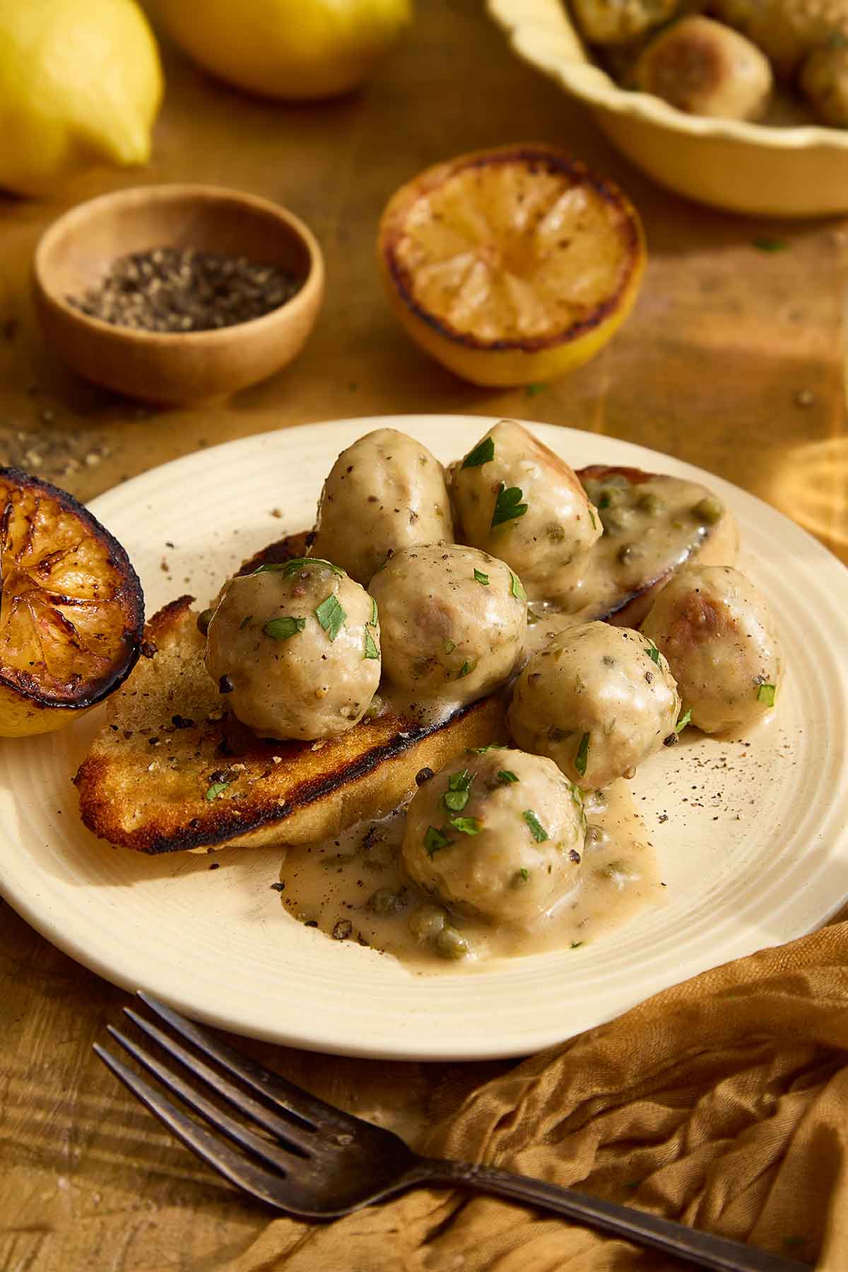 Close-up of chicken piccata meatballs over grilled bread with charred lemon and glossy lemon-butter sauce.