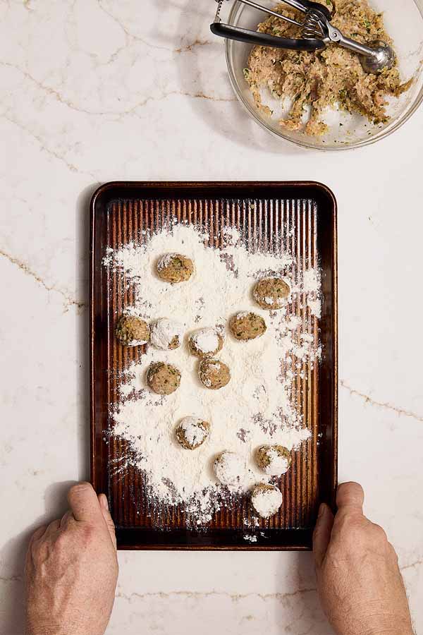 Chicken meatballs being rolled in flour on a tray for pan-frying.