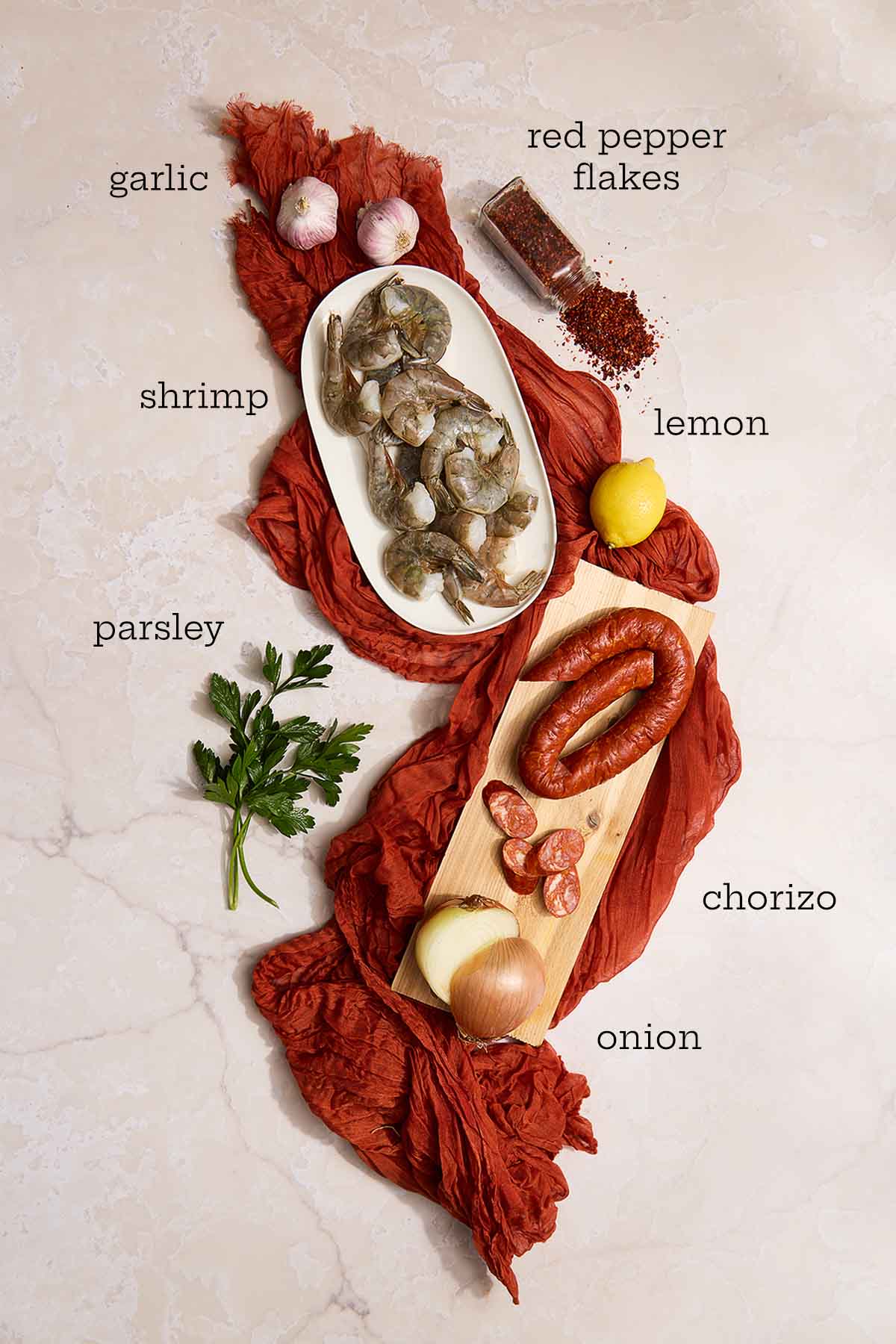 Raw shrimp, chorizo, onion, garlic, parsley, lemon, and red pepper flakes arranged on a light background.