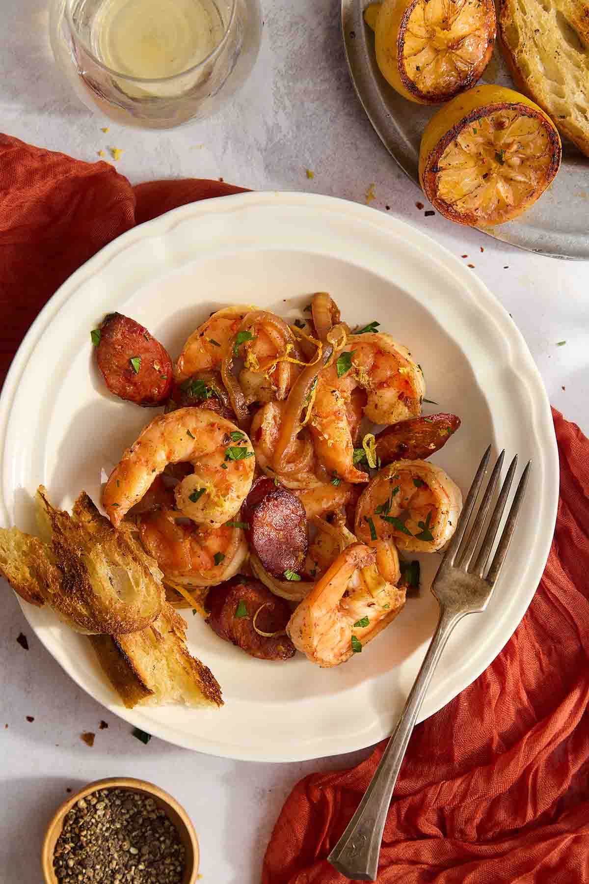 Overhead view of shrimp and chorizo in a white bowl, topped with parsley and lemon zest, served with grilled lemon and toasted bread.