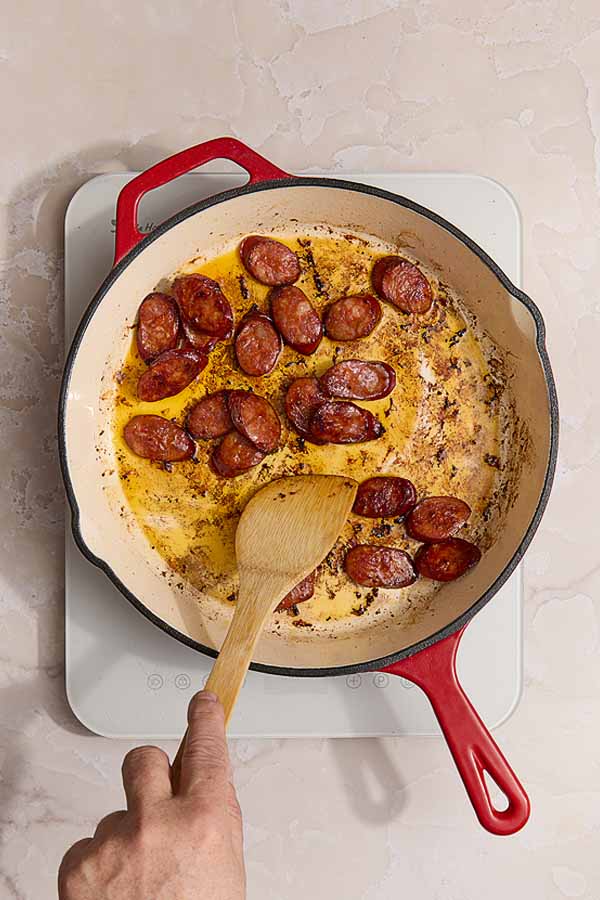Sliced chorizo sizzling in a red-handled skillet, beginning to brown and release oil.