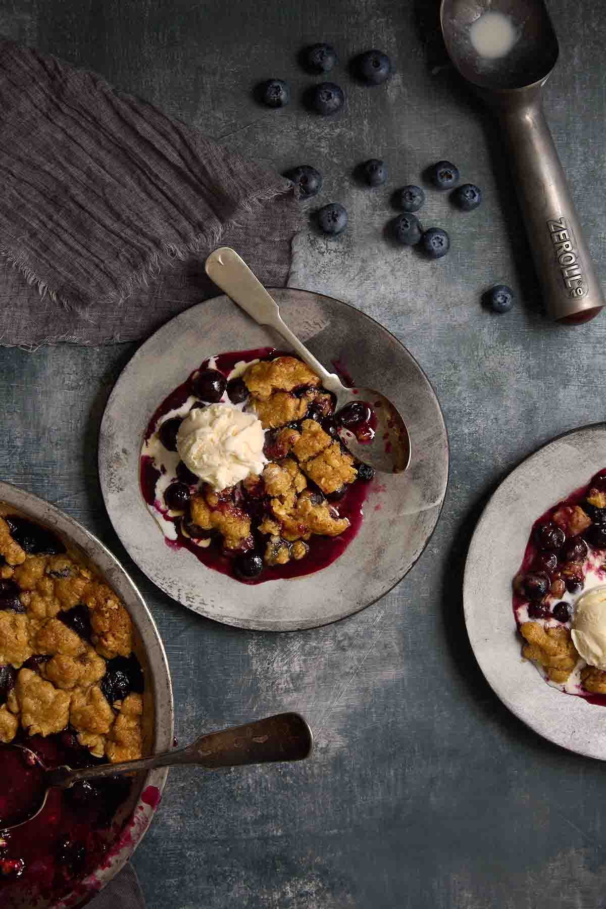 Blueberry crumble with melted vanilla ice cream and blueberries on a grey plate.