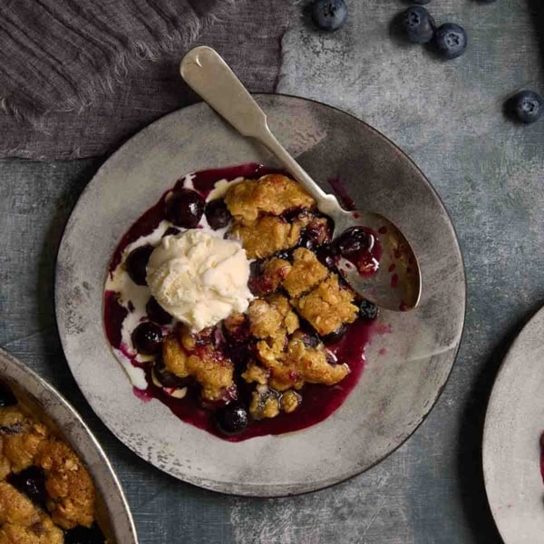 A square image of blueberry crumble with a scoop of vanilla ice cream.