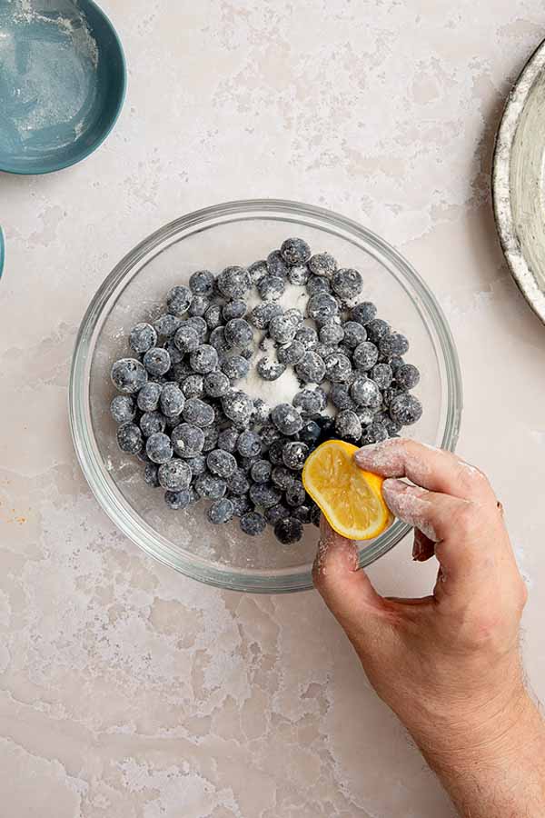 Tossing fresh blueberries with sugar and lemon juice in a clear bowl.