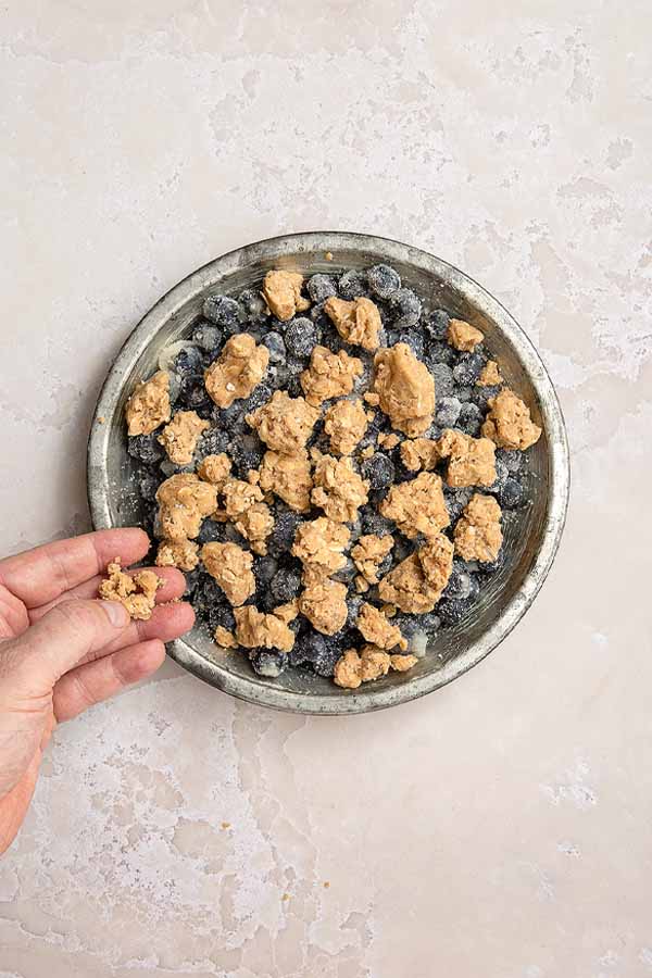 Adding the prepared crumble topping in clumps over the blueberries in the pie pan.