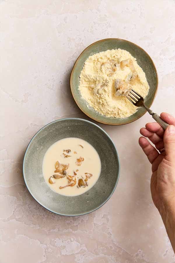 A hand using a fork to lift a clam from a bowl of buttermilk into a bowl of corn flour and cake flour, for fried clams.