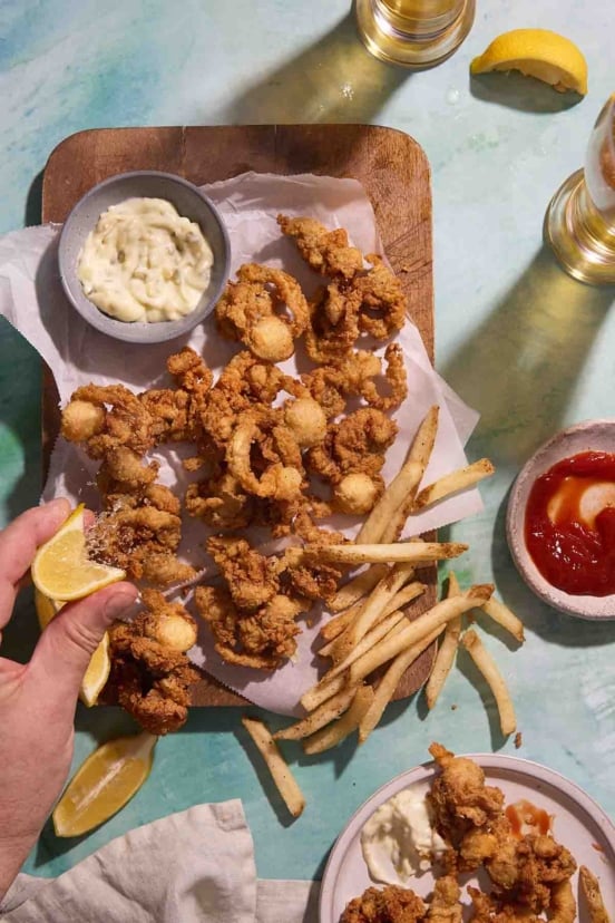 A hand squeezing lemon over a platter of crispy fried whole belly clams and French fries, with bowls of tartar sauce and ketchup.