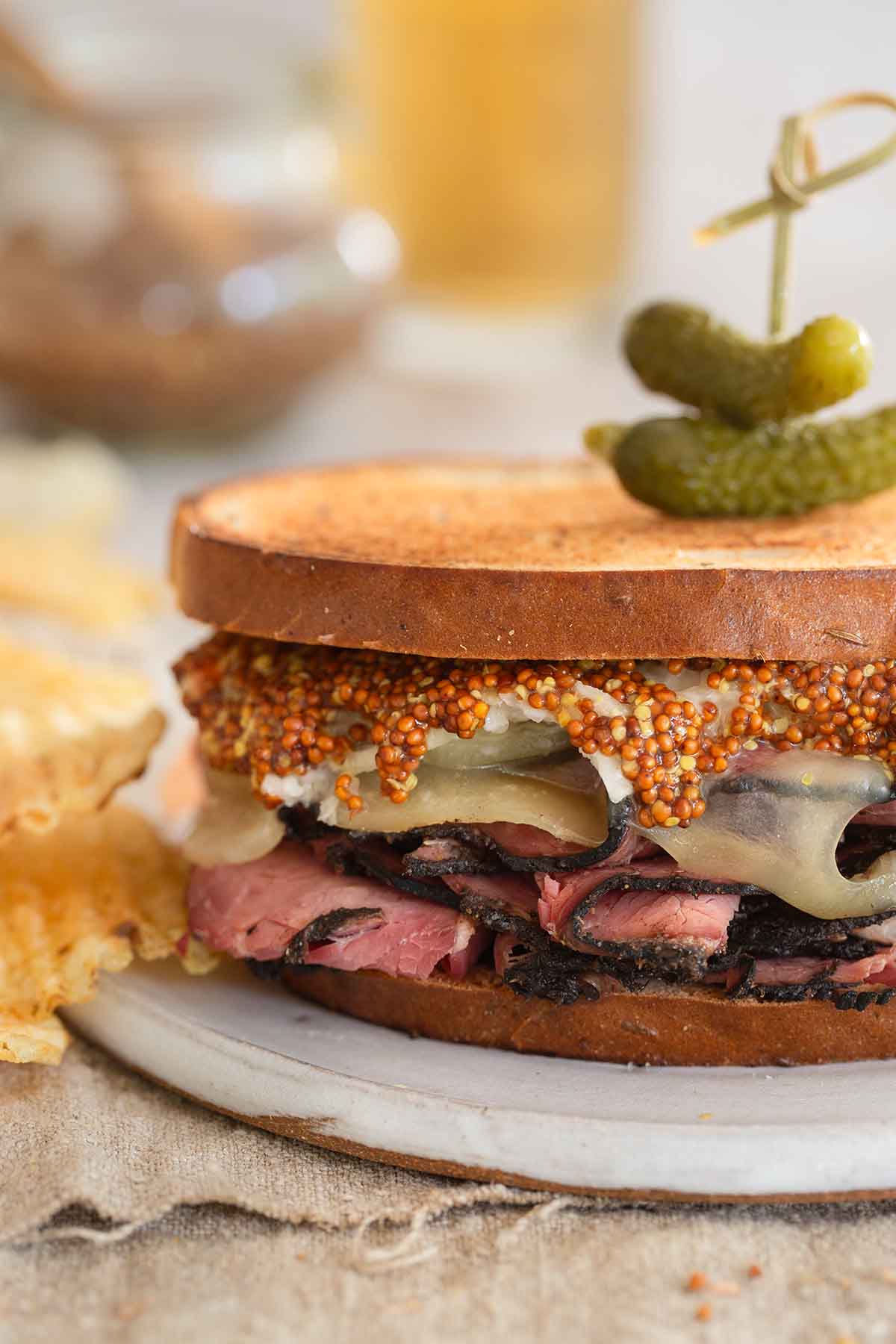 A homemade pastrami sandwich with two small pickles on top, served with a side of potato chips.