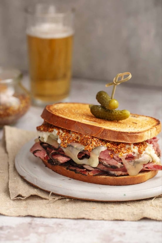 A homemade pastrami sandwich on toasted rye bread with melted cheese, whole grain mustard, and small pickles on top.