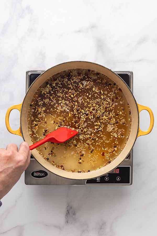 A hand stirring a large yellow pot of brown brining liquid with whole spices on an induction burner.