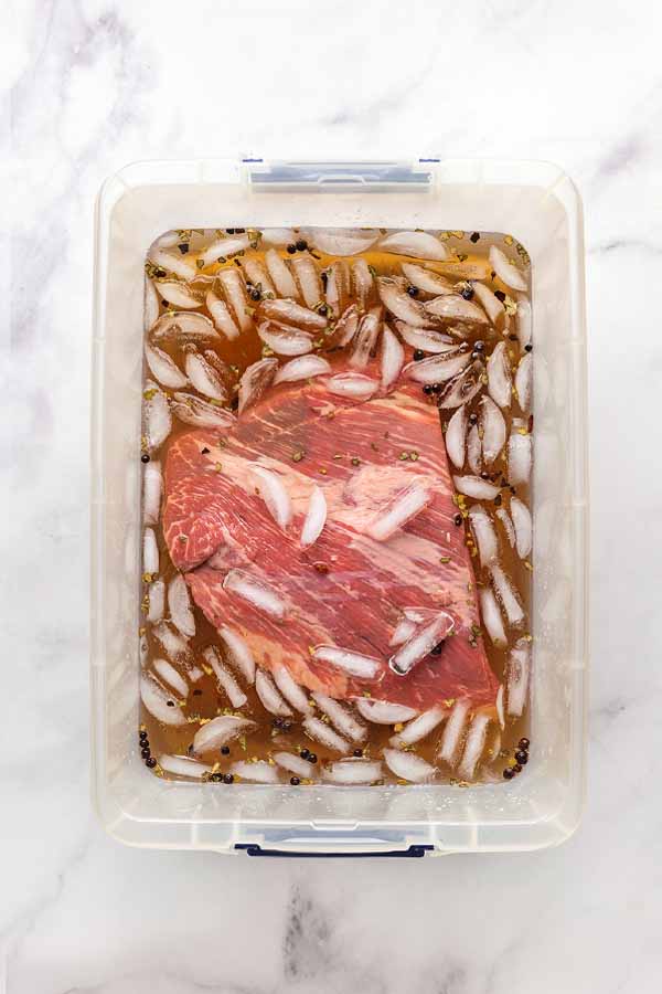 A piece of beef brisket submerged in a clear plastic container filled with brining liquid and ice.