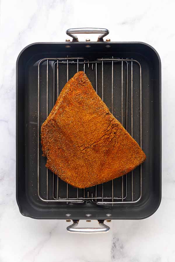 A spice-rubbed beef brisket resting on a wire rack inside a black roasting pan.