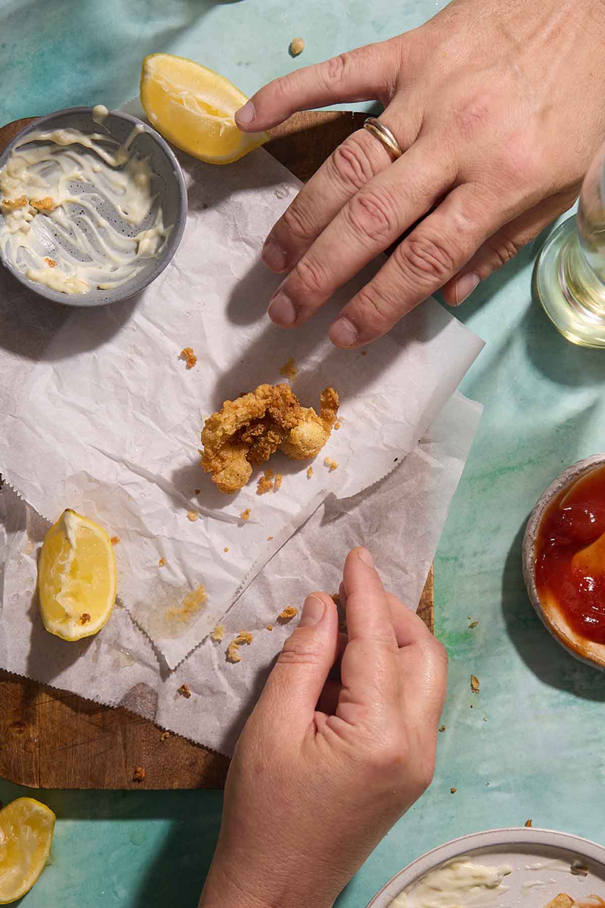 single crispy fried clam on parchment paper, with hands reaching for it and bowls of tartar sauce and ketchup nearby.