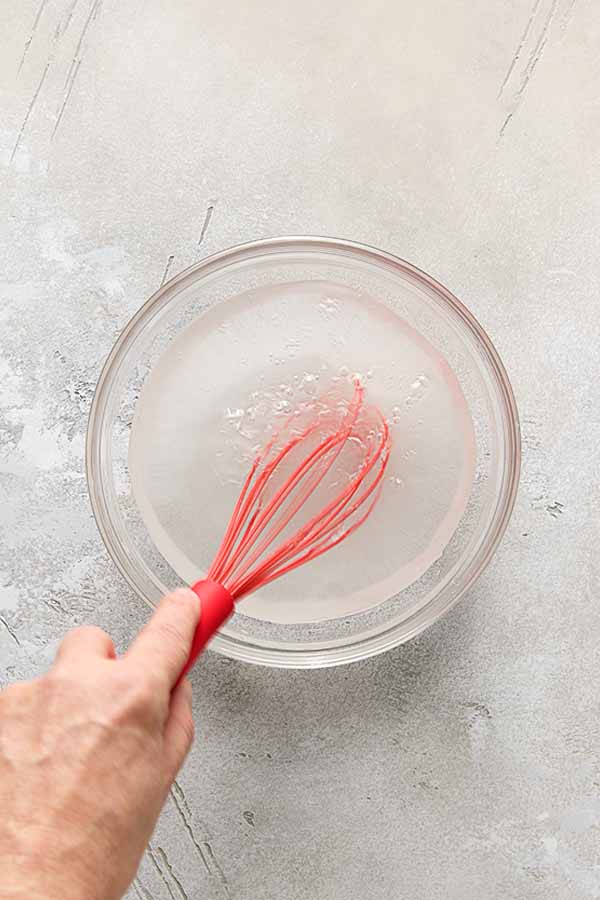 A hand whisking salt and sugar into water in a glass bowl to make a brine for fried chicken.