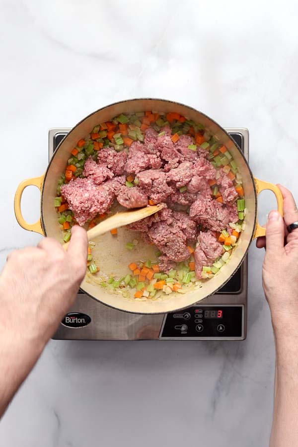 Ground beef, veal, and pork browning in a Dutch oven, being stirred with a wooden spoon.