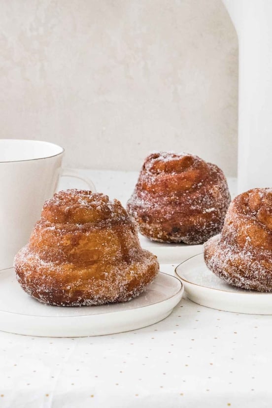 Three morning buns on small white plates, dusted with granulated sugar, with a coffee cup behind them.