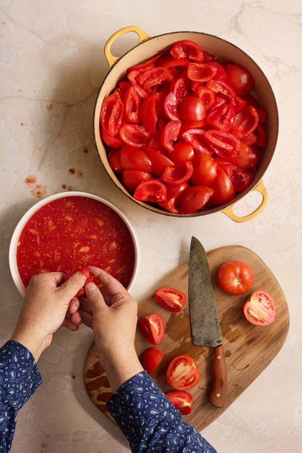 Hands removing seeds from quartered tomatoes over a white bowl, with a pot of seeded tomatoes nearby.
