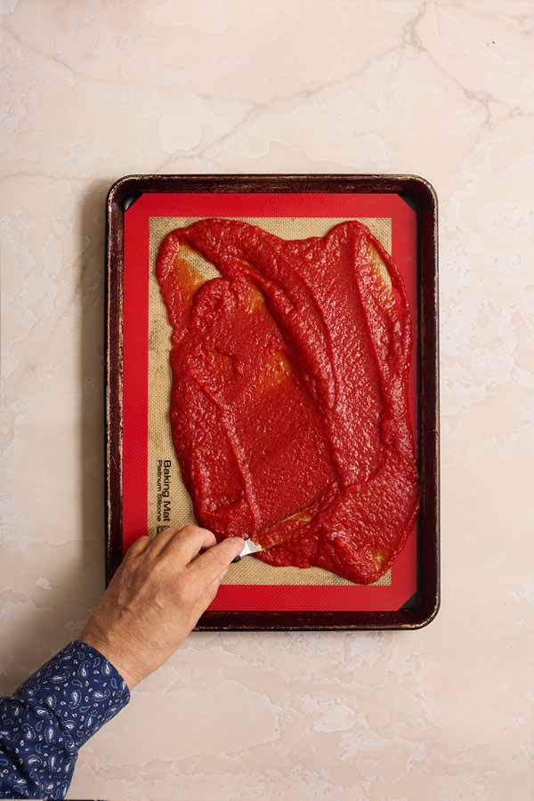 A hand using a small spatula to spread thick tomato purée onto a silicone mat on a baking sheet.
