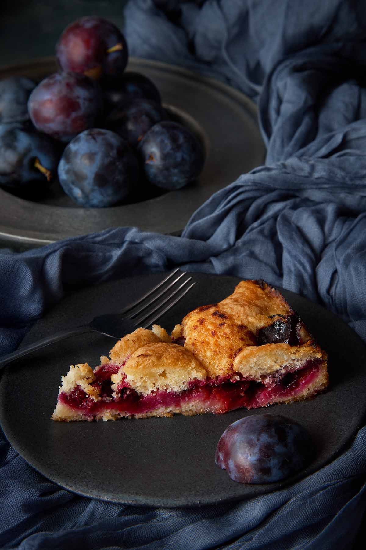 A slice of classic plum torte with a jammy fruit layer on a dark plate.