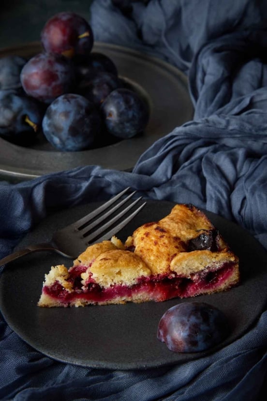 A slice of classic plum torte with a jammy fruit layer on a dark plate.