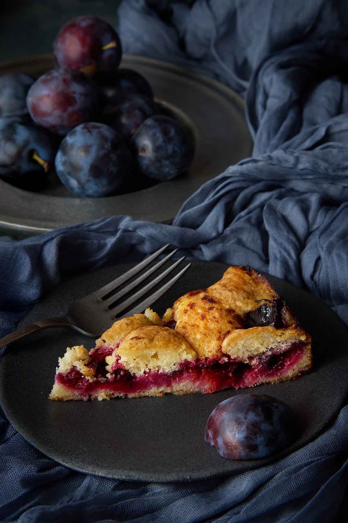 A slice of classic plum torte with a jammy fruit layer on a dark plate.