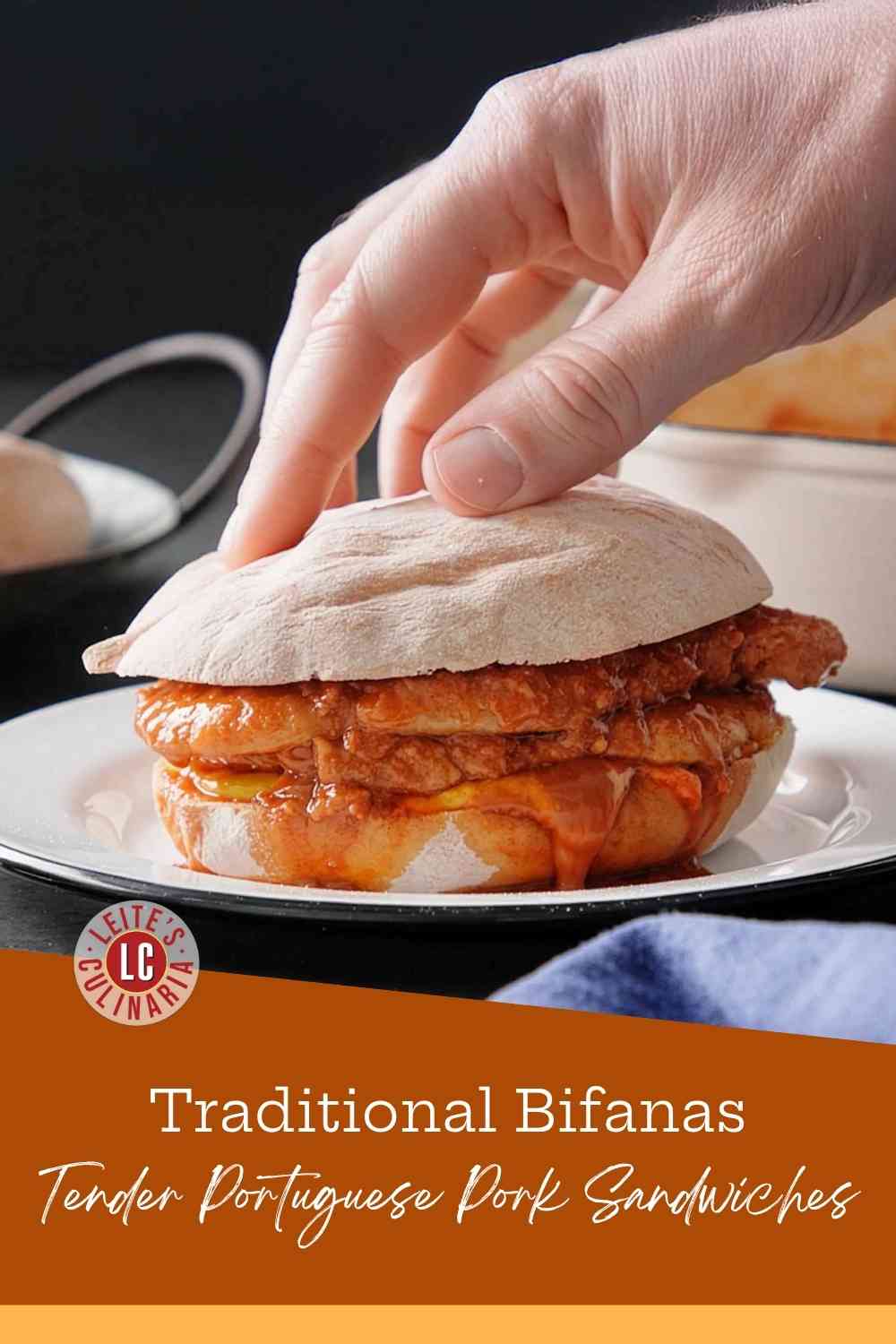 A man's hand pressing down on a Portuguese roll filled with thin pork slices in a paprika-white wine sauce, with mustard and piri-piri sauce dripping.