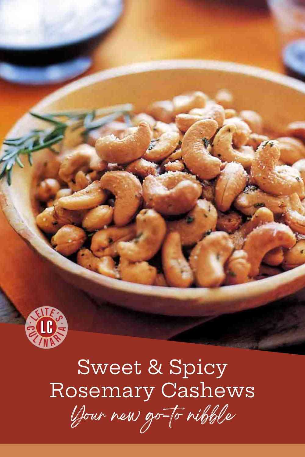 A wooden bowl overflowing with golden-brown roasted cashews seasoned with rosemary and salt, with a fresh herb sprig.