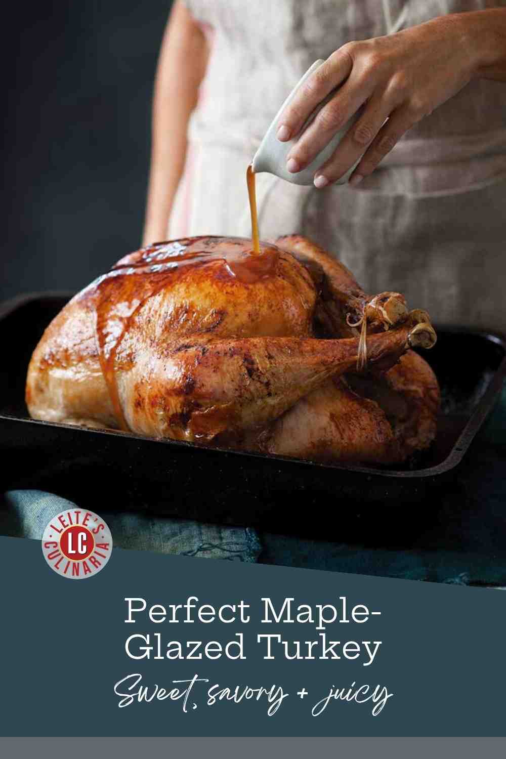 A glossy, mahogany-brown maple-glazed turkey resting in a black roasting pan as a cook pours on a final layer of glaze.