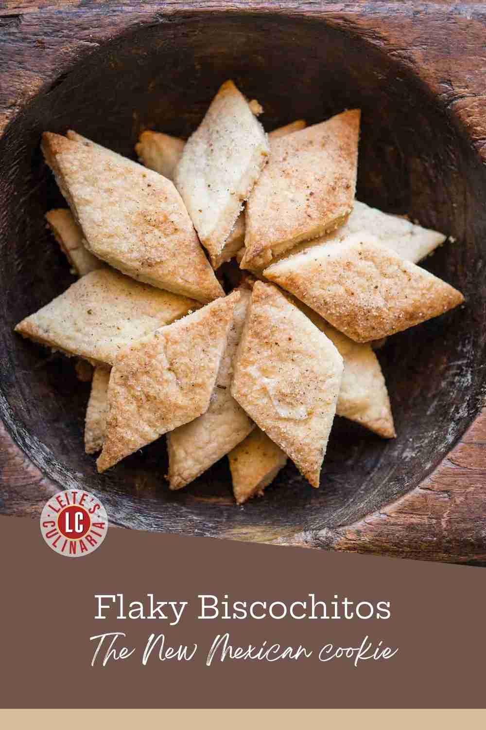 A cluster of diamond-shaped, sugar-dusted biscochitos, flaky anise-flavored shortbread, nestled in a dark wooden serving bowl.