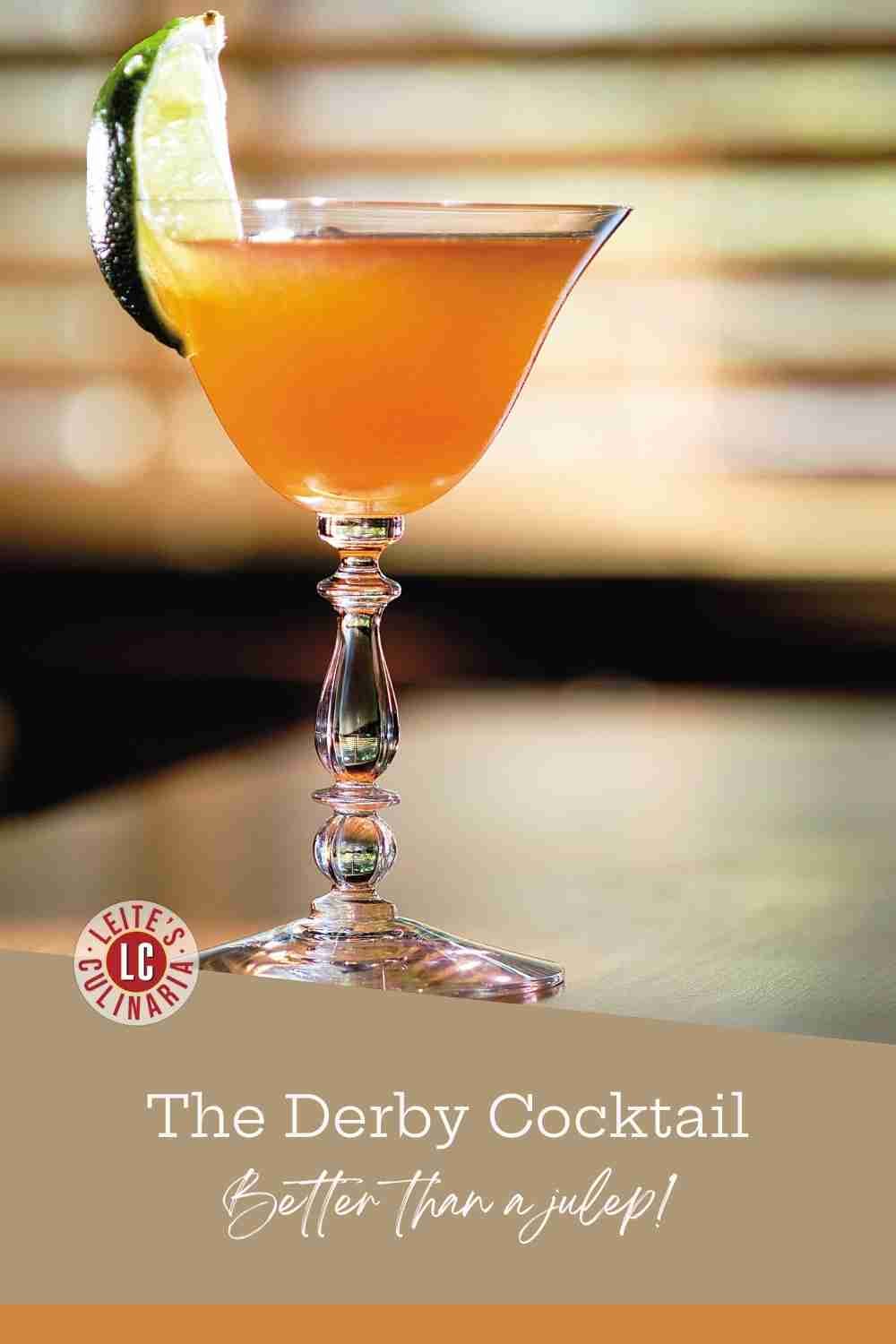 A golden-amber cocktail in a stemmed glass garnished with a lime wedge against a blurred background.