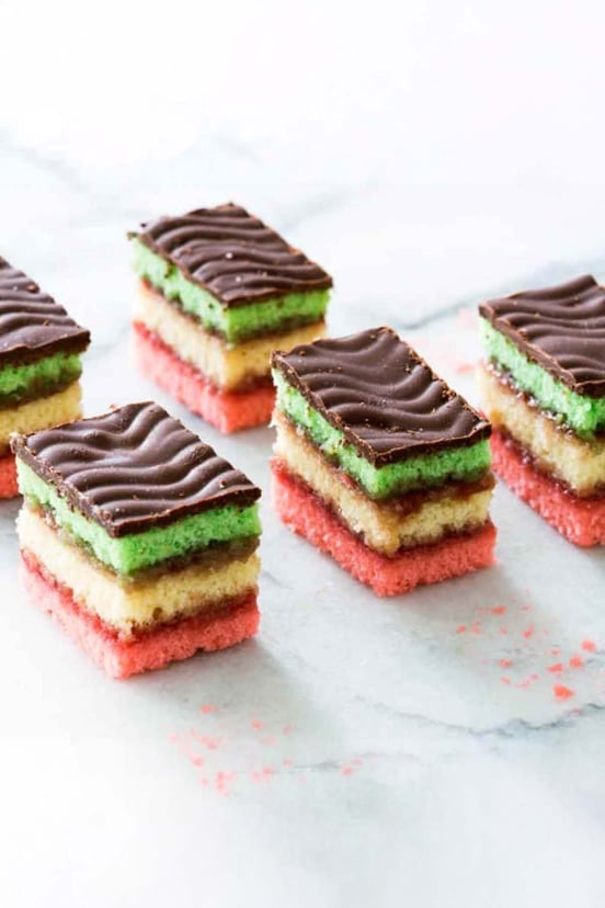 Five squares of Italian rainbow cookies, with layers of pink, yellow, green cake layers and a chocolate topping.