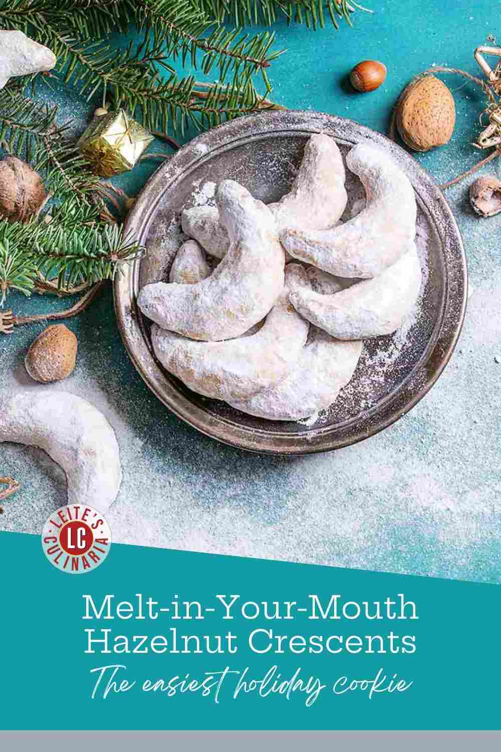 Powder-dusted hazelnut crescent cookies on a silver platter with pine sprigs, jazelnuts, and gold ornaments.