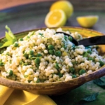 Vibrant green peas and yellow lemon zest scattered through fluffy pearl couscous in a sunny, rustic ceramic bowl.