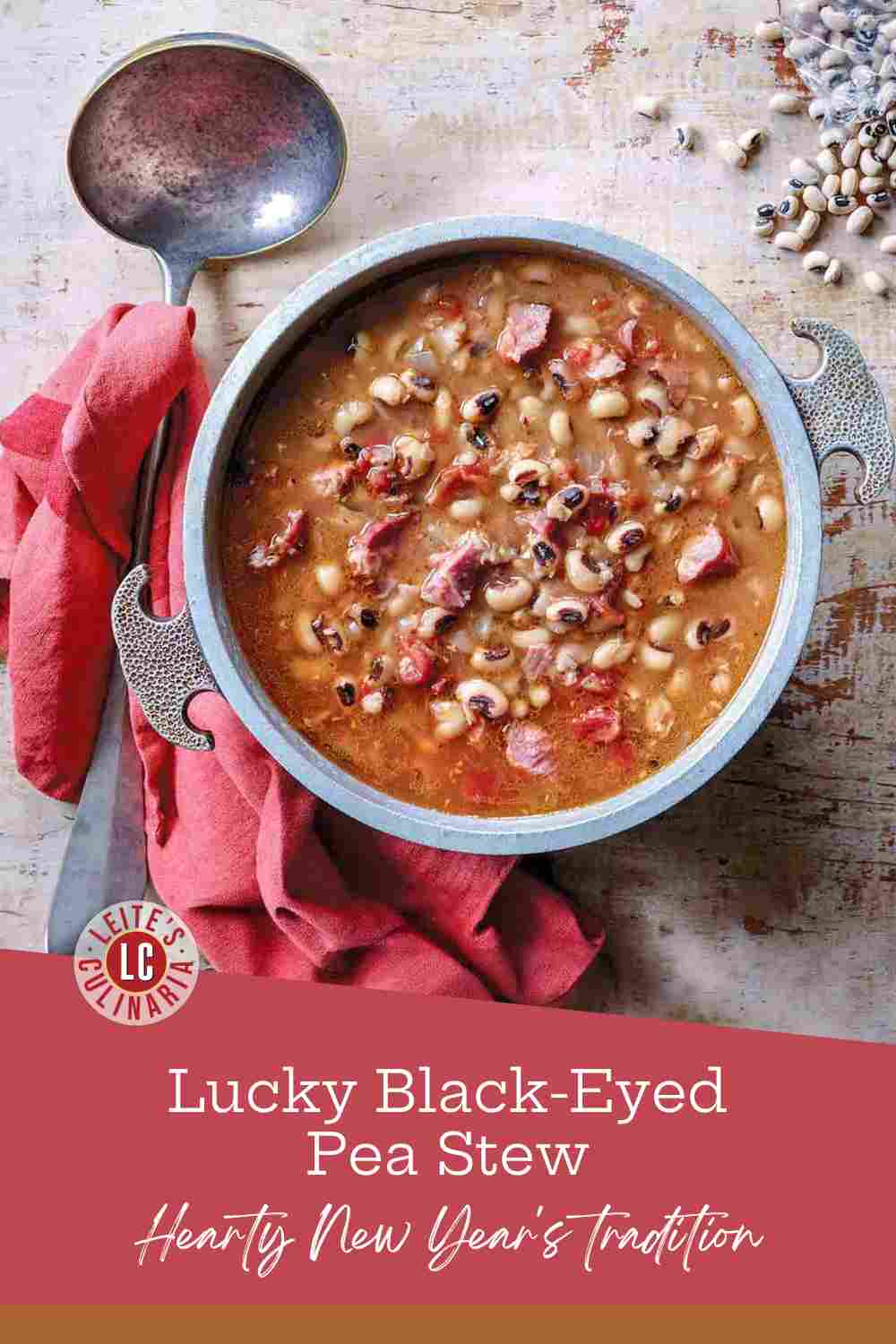Hearty black-eyed pea stew with chunks of ham in a silver pot next to a red napkin.