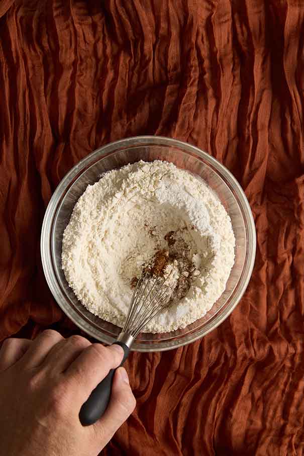 Hand whisking flour, baking powder, and nutmeg in a glass bowl over a rustic cloth.