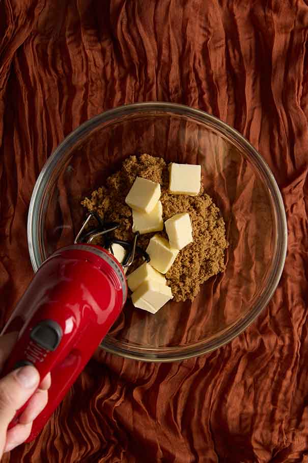 Red hand mixer beating cubes of butter and brown sugar in a glass mixing bowl.