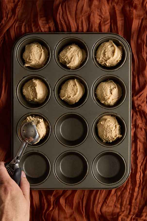 A hand using a cookie scoop to portion thick muffin batter into a dark 12-cup muffin tin.