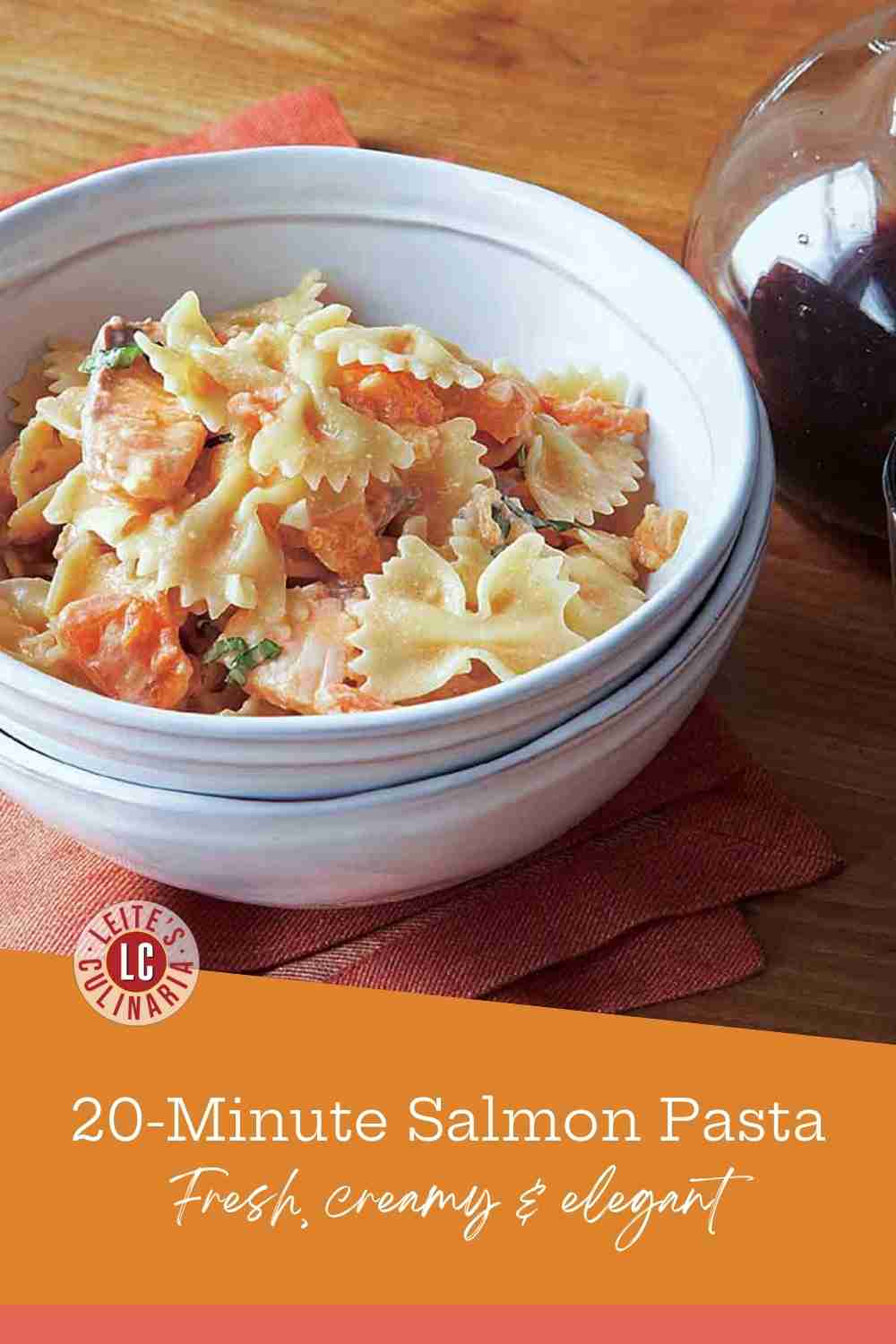 A white bowl of bow tie pasta with chunks of pink salmon in a light tomato cream sauce on a wooden table.