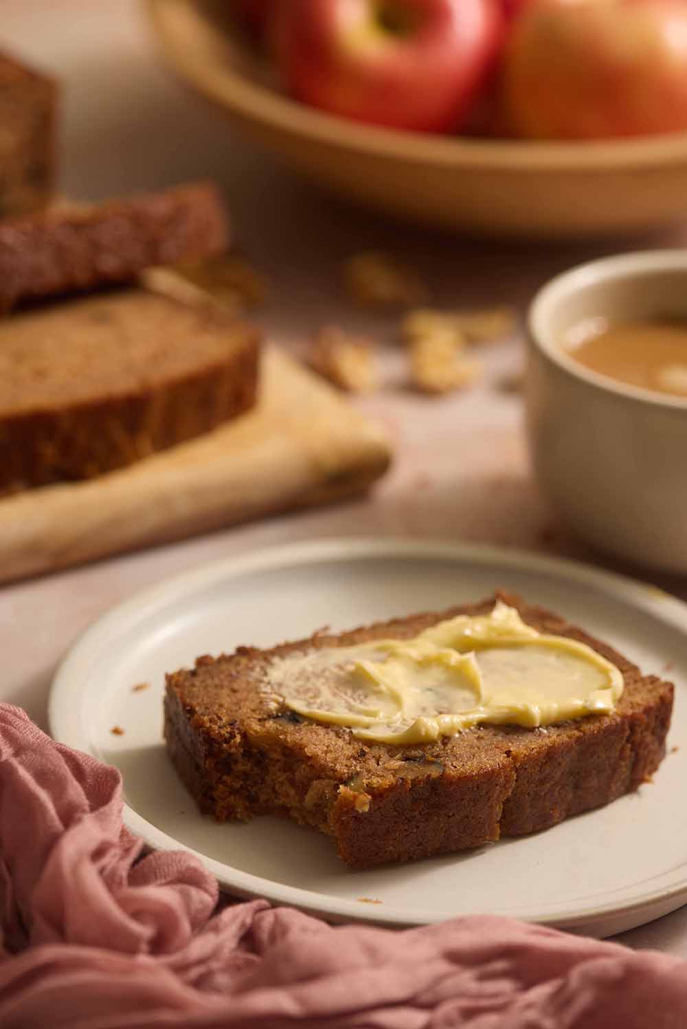 A slice of moist applesauce bread on a tan plate with a cup of coffee and a bowl of apples behind it.