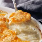 A spoon of golden-brown scalloped potatoes being scooped from a white casserole dish.