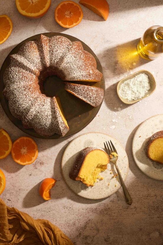 Whole Portuguese orange olive oil Bundt cake dusted with sugar on a wooden table with orange slices.