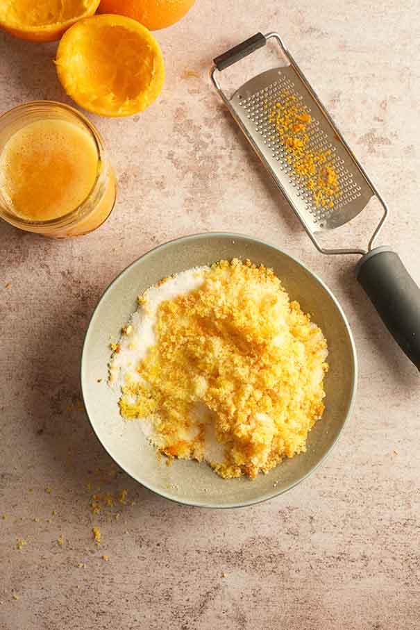 Grated orange zest being rubbed into white granulated sugar in a small ceramic bowl.