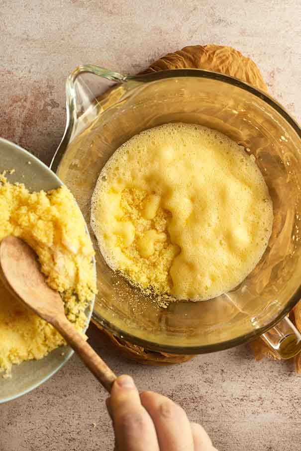Orange sugar being poured into a glass bowl of beaten eggs to create a thick cake batter.