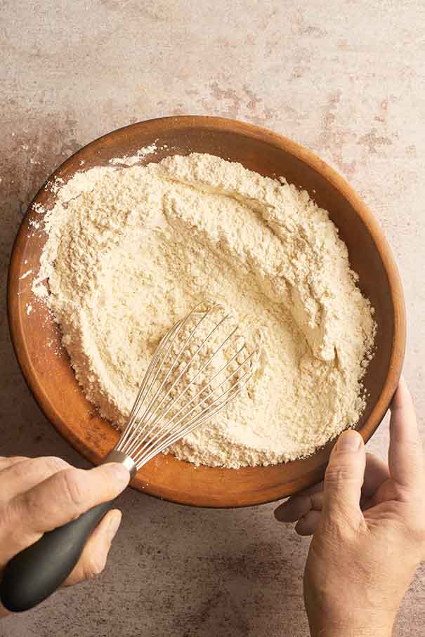 A metal whisk mixing all-purpose flour, baking powder, and salt in a large wooden bowl.