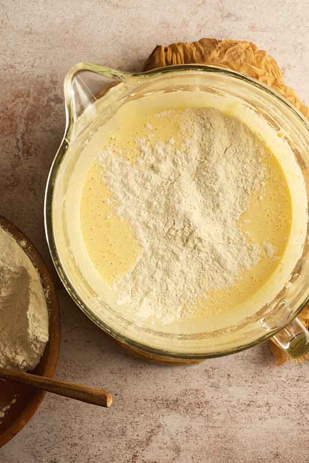 Sifted flour mixture being added to the pale yellow egg and sugar base in a glass mixing bowl.