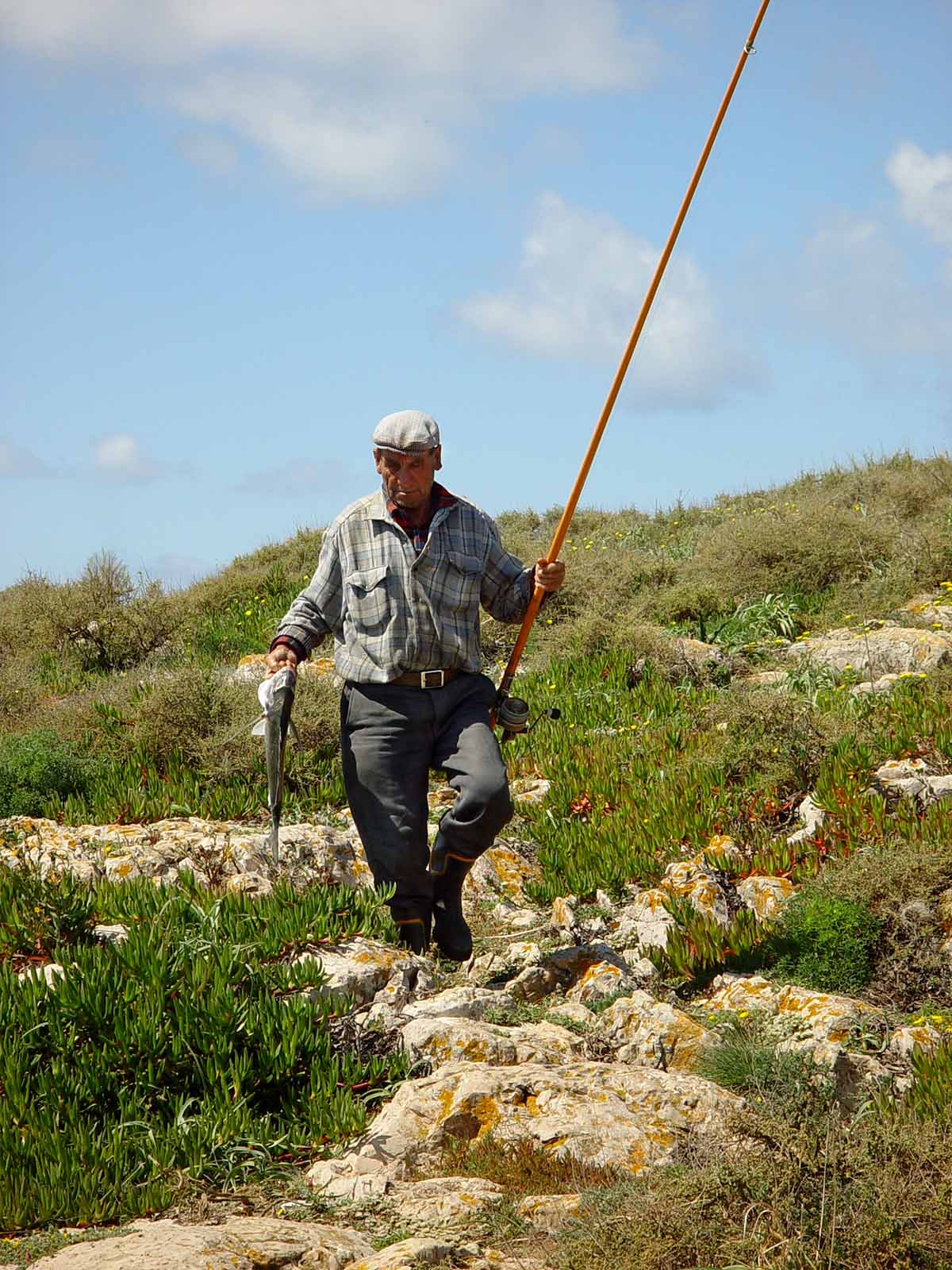 A Portuguese fisherman, walking down a rocky hill holding a fish he just caught.