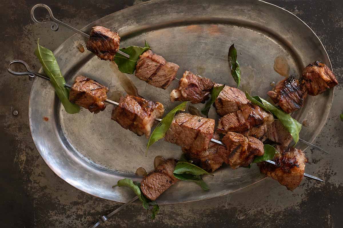 Three metal skewers and chunks of grilled beef and bay leaves on a pewter plate.