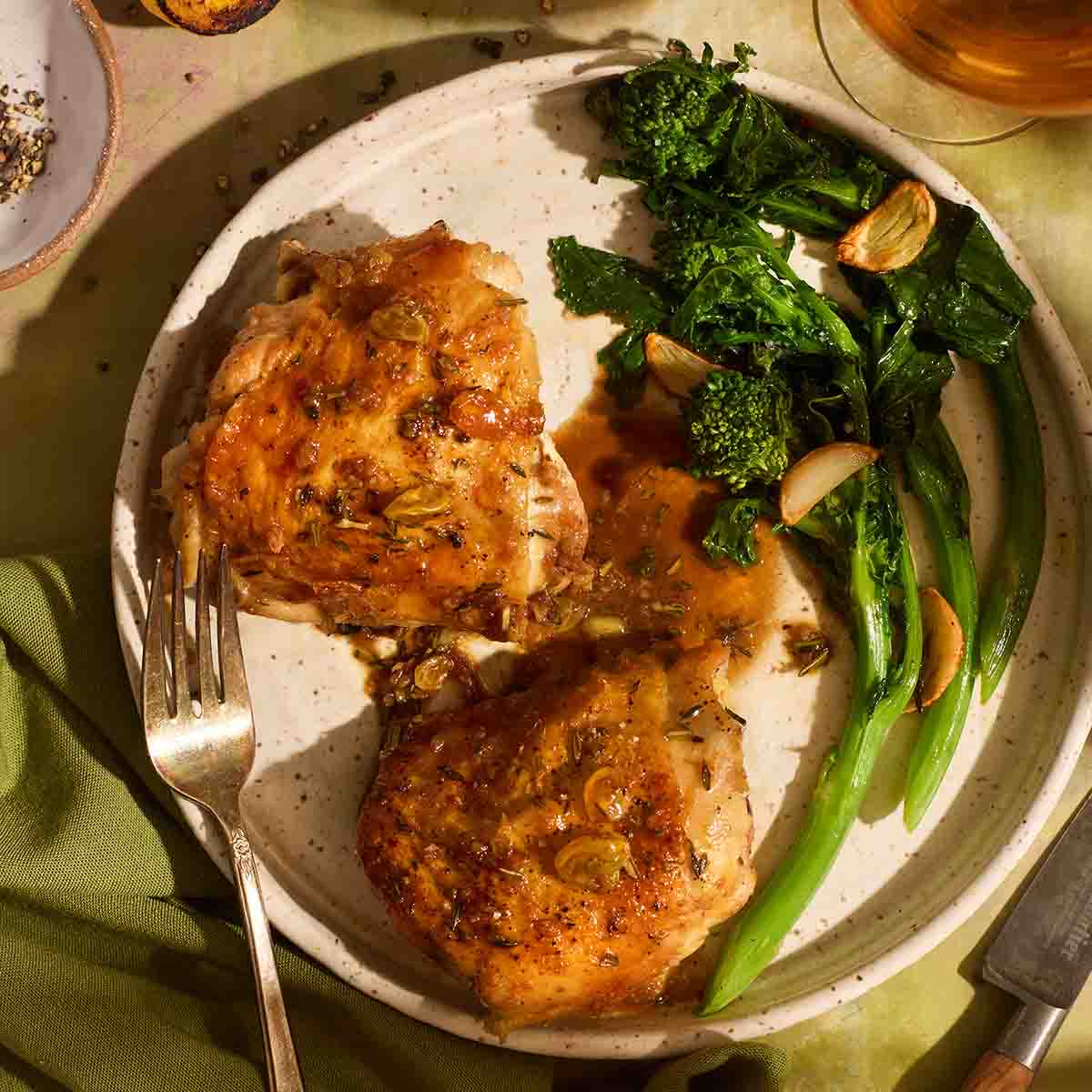 Close up of pan-seared chicken thighs drizzled with an agrodolce sauce featuring golden raisins, plated alongside roasted green broccolini.
