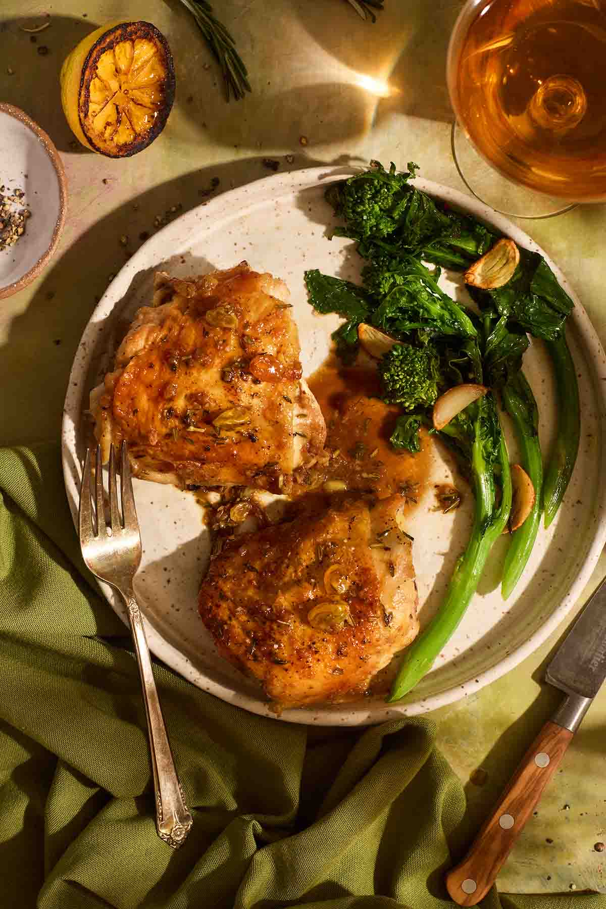 Two chicken thighs topped with golden raisin sweet and sour sauce, served on a speckled plate with vibrant green broccolini, a charred lemon half, and a glass of white wine.